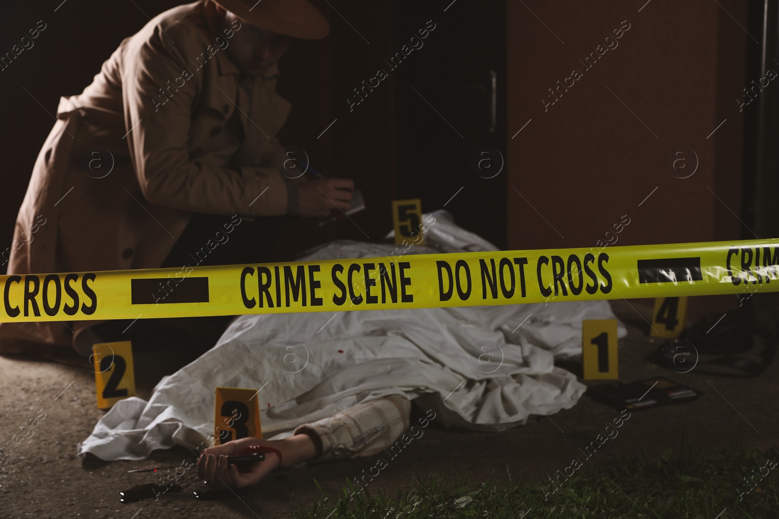 Photo of Investigator examining crime scene with dead body outdoors, focus on yellow tape