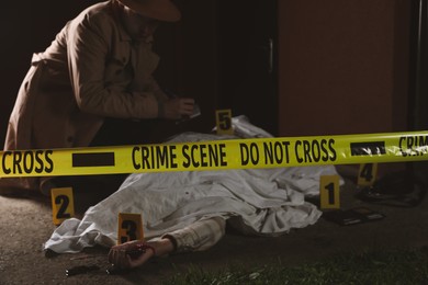 Photo of Investigator examining crime scene with dead body outdoors, focus on yellow tape