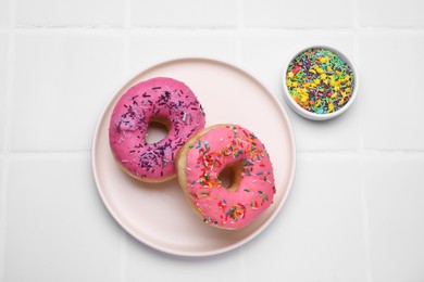 Photo of Glazed donuts decorated with sprinkles on white tiled table, top view. Tasty confectionery