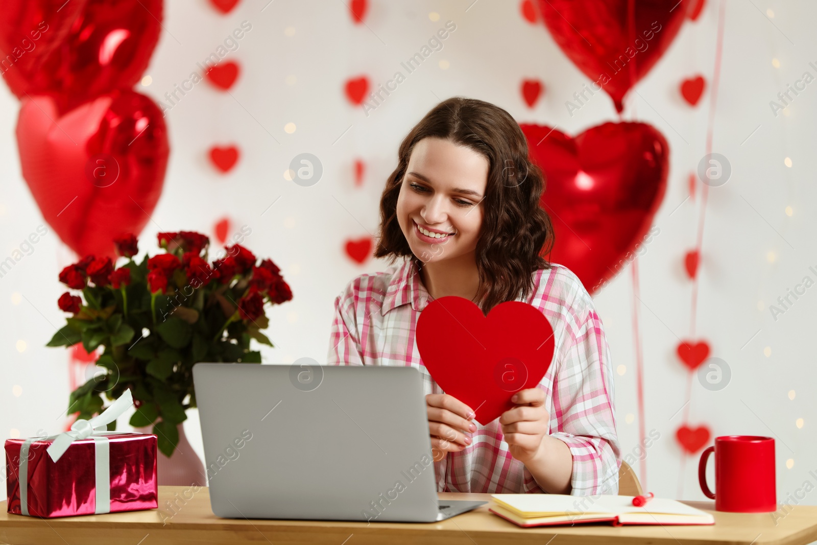 Photo of Valentine's day celebration in long distance relationship. Woman holding red paper heart while having video chat with her boyfriend via laptop at home