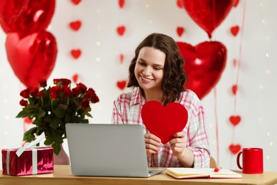 Photo of Valentine's day celebration in long distance relationship. Woman holding red paper heart while having video chat with her boyfriend via laptop at home