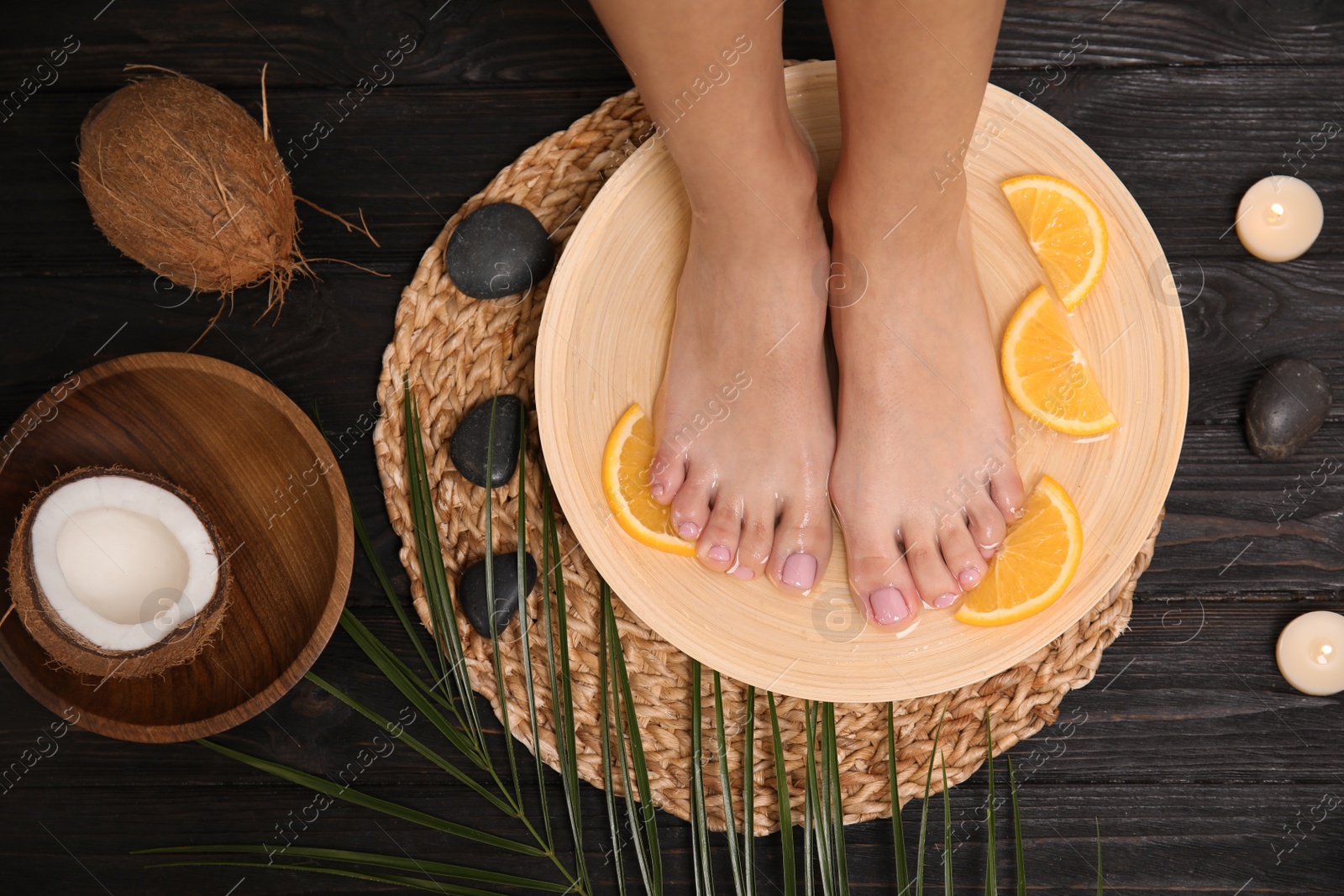 Photo of Woman soaking her feet in plate with water and orange slices on wooden floor, top view. Spa treatment