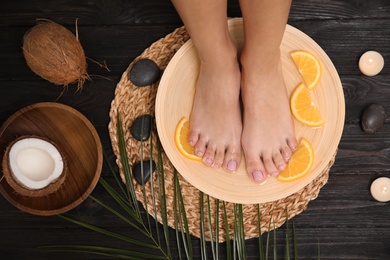 Woman soaking her feet in plate with water and orange slices on wooden floor, top view. Spa treatment