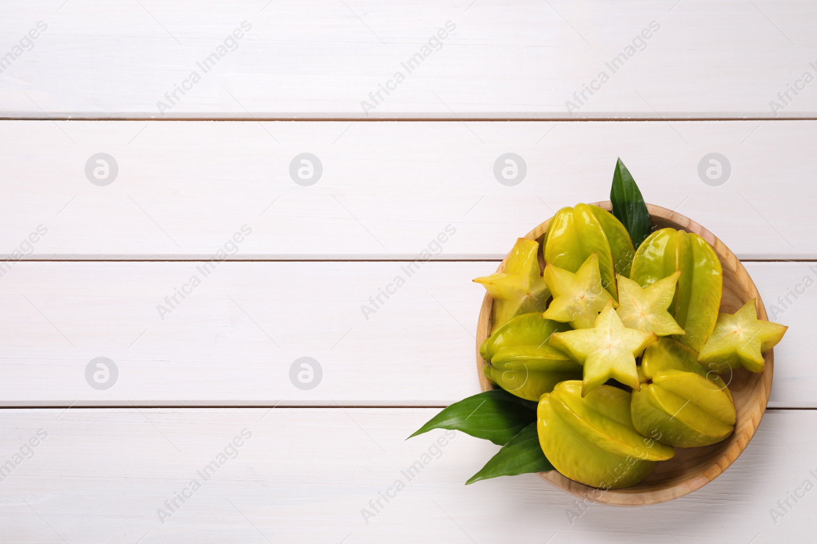 Photo of Cut and whole carambolas with green leaves in bowl on white wooden table, top view. Space for text