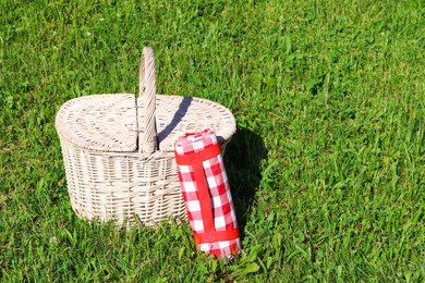 Photo of Rolled checkered tablecloth near picnic basket on green grass outdoors, space for text