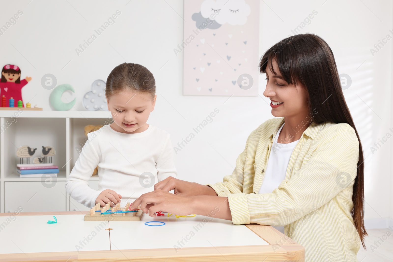 Photo of Motor skills development. Mother helping her daughter to play with geoboard and rubber bands at white table in room