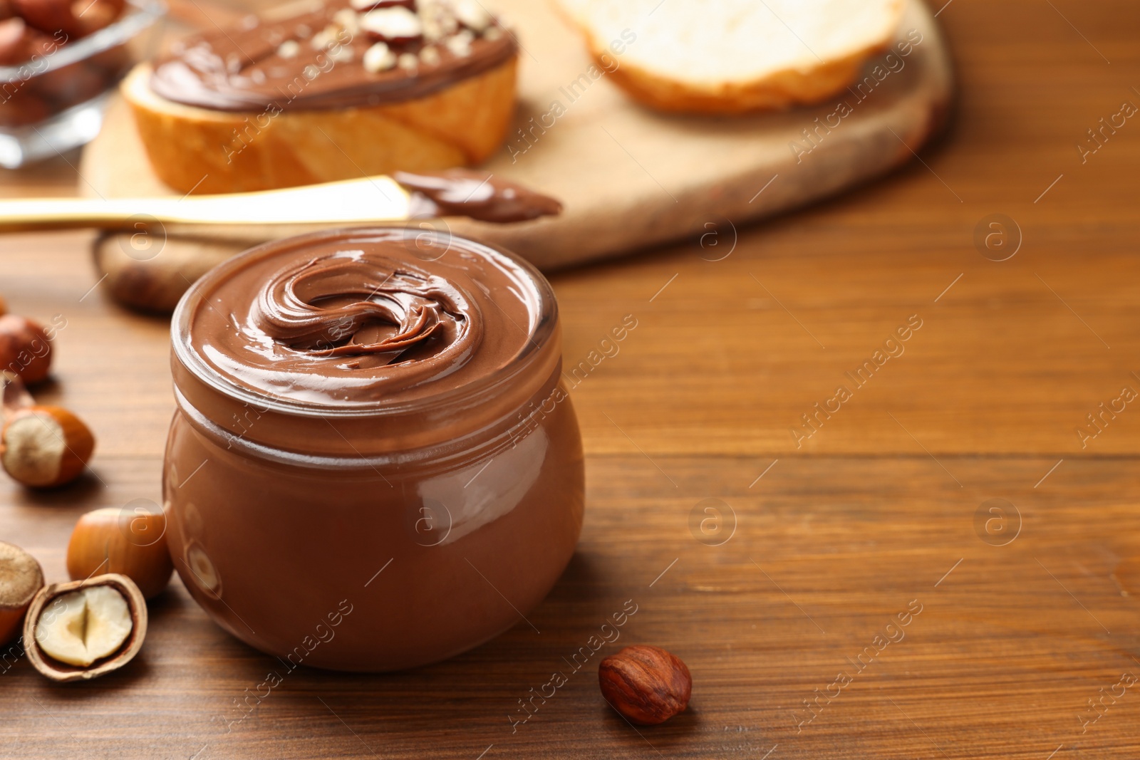Photo of Glass jar with tasty chocolate hazelnut spread and nuts on wooden table. Space for text