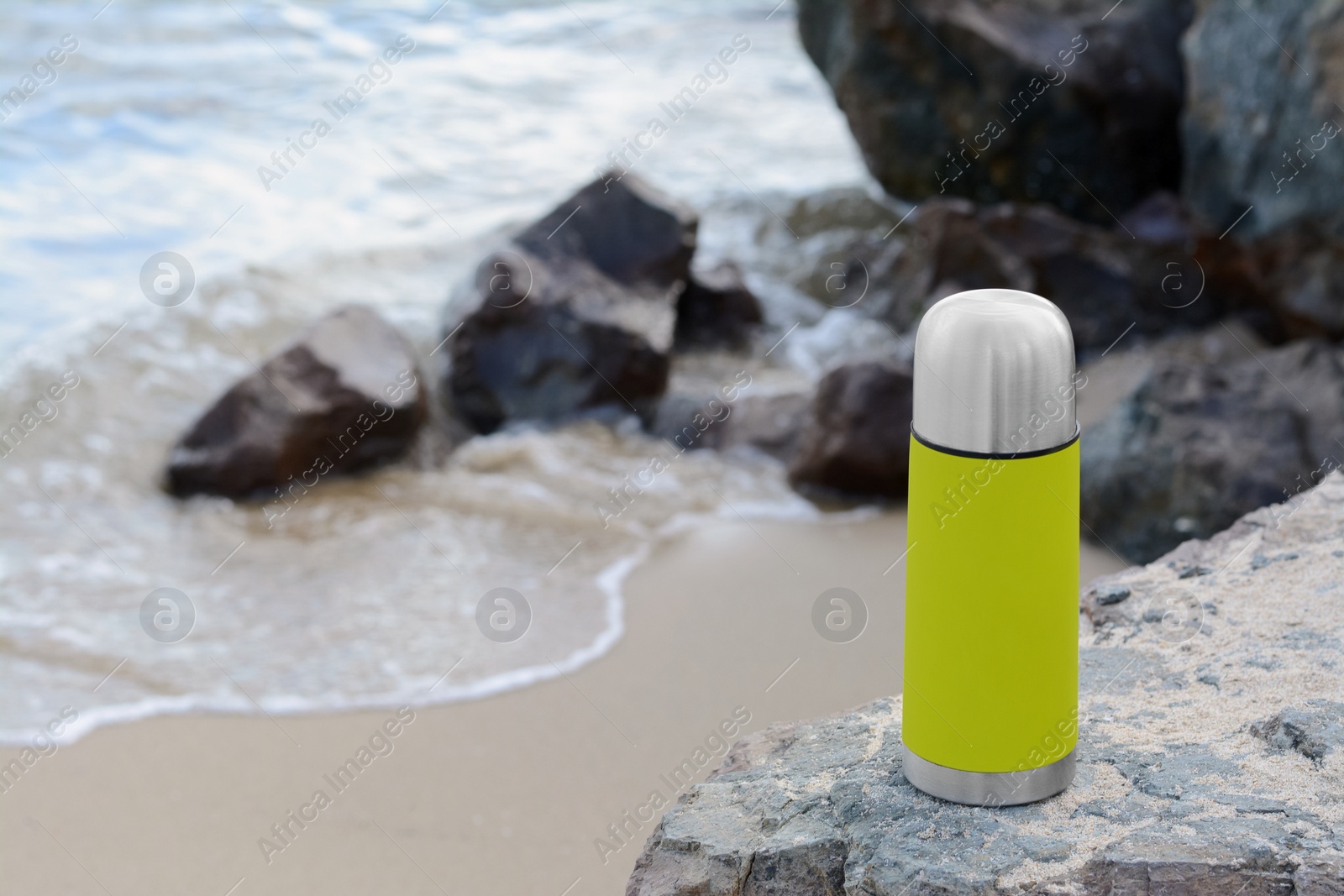 Photo of Metallic thermos with hot drink on stone near sea, space for text