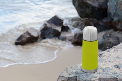 Metallic thermos with hot drink on stone near sea, space for text