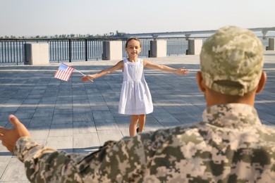 Photo of Little girl with flag of USA running towards her father in military uniform outdoors. Family reunion