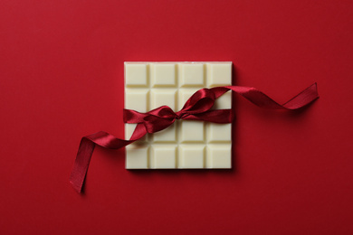 Photo of Tasty white chocolate bar with ribbon on red background, top view