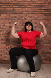 Photo of Happy overweight mature woman sitting on fitness ball near brick wall indoors