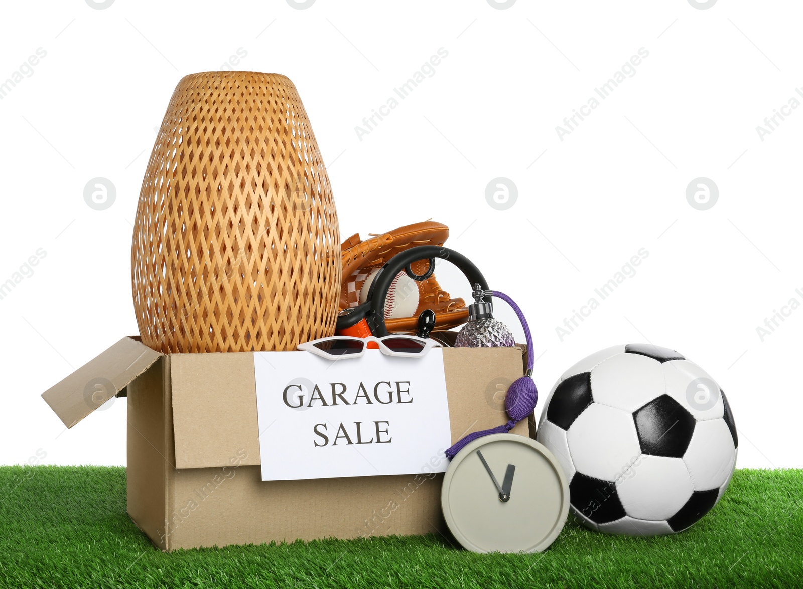 Photo of Box with sign Garage Sale and different stuff on green grass against white background