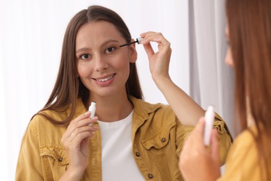 Photo of Beautiful woman applying serum onto eyelashes near mirror indoors