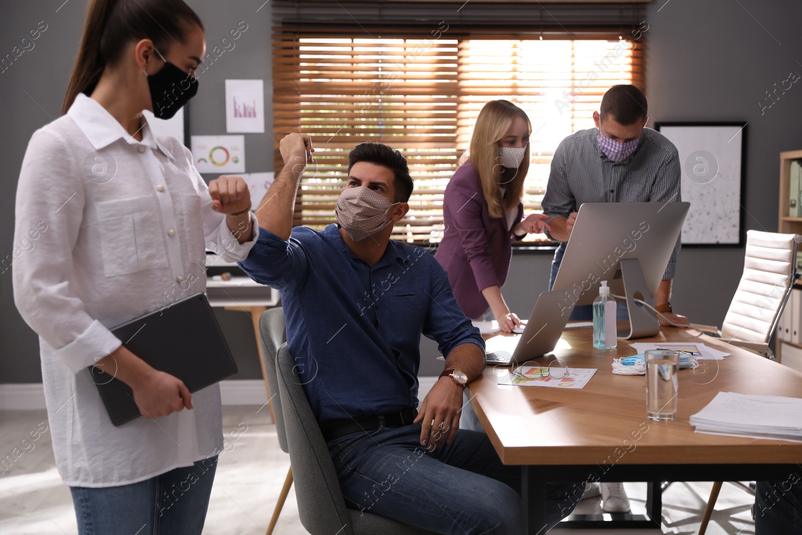 Photo of Coworkers with protective masks making elbow bump in office. Informal greeting during COVID-19 pandemic