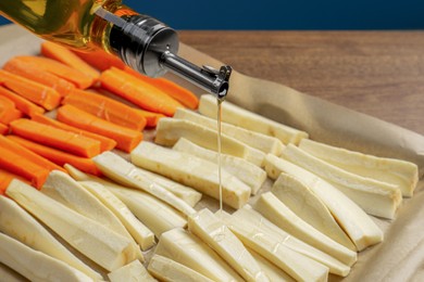 Pouring oil onto baking tray with parsnips and carrots against blue background, closeup