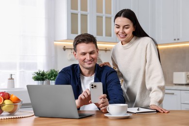 Photo of Happy couple using smartphone together at wooden table in kitchen