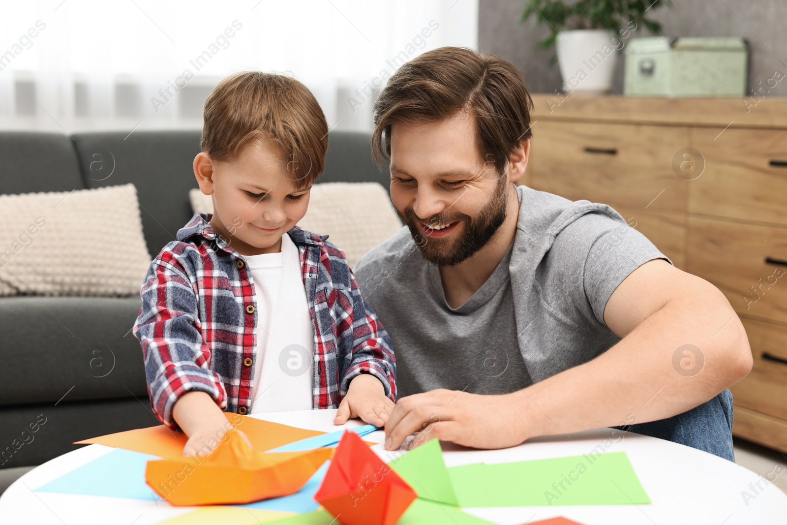 Photo of Dad and son making paper boats at coffee table indoors