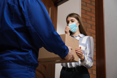 Photo of Young woman wearing medical mask receiving parcel from delivery man indoors. Prevention of virus spread