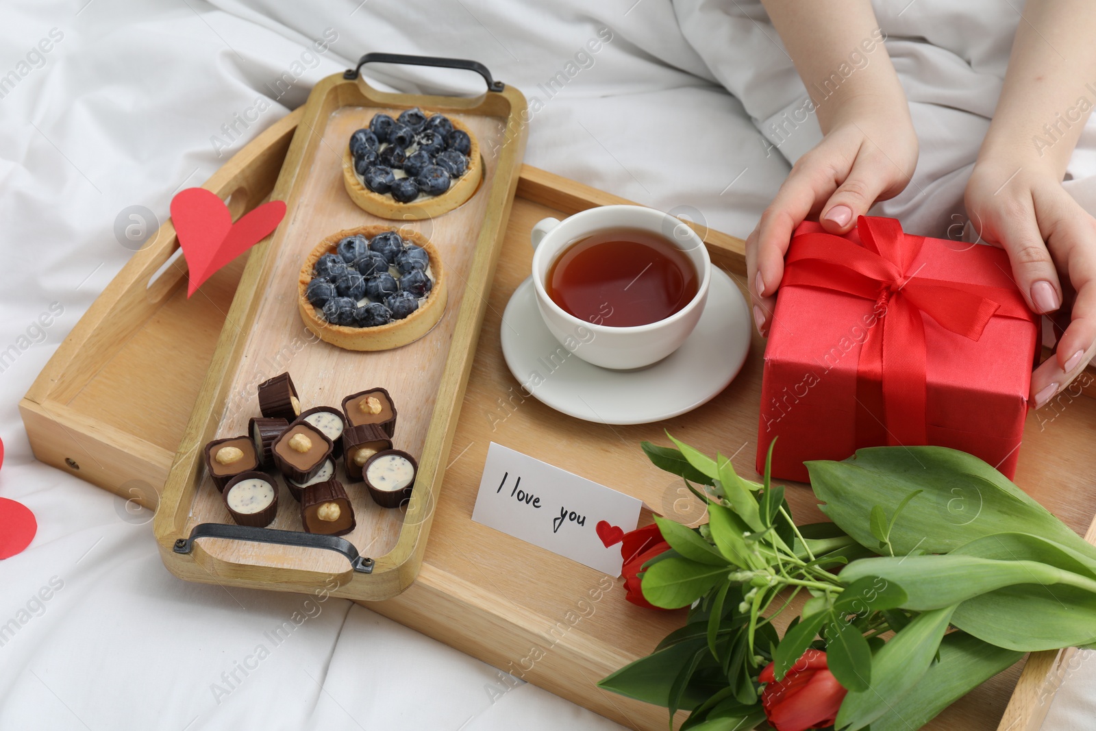 Photo of Tasty breakfast served in bed. Woman with gift box, tea, desserts, flowers and I Love You card at home, closeup