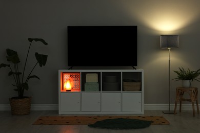 Modern TV on cabinet, lamp and beautiful houseplants near light wall indoors. Interior design