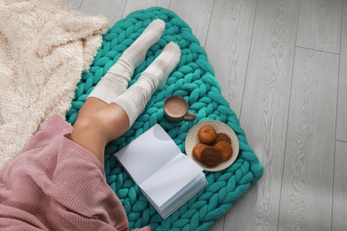 Photo of Young woman with book, cookies and cup of coffee on knitted blanket at home, top view. Winter atmosphere