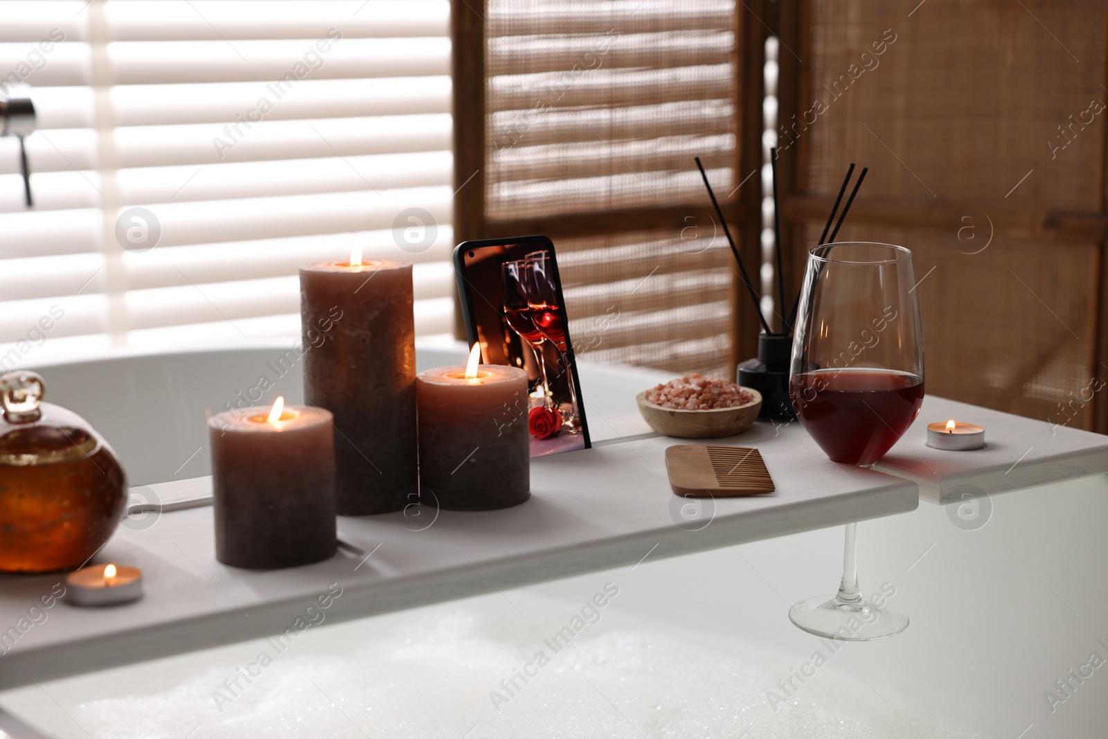 Photo of White wooden tray with smartphone, glass of wine and burning candles on bathtub in bathroom