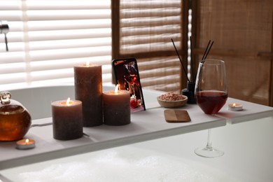 Photo of White wooden tray with smartphone, glass of wine and burning candles on bathtub in bathroom