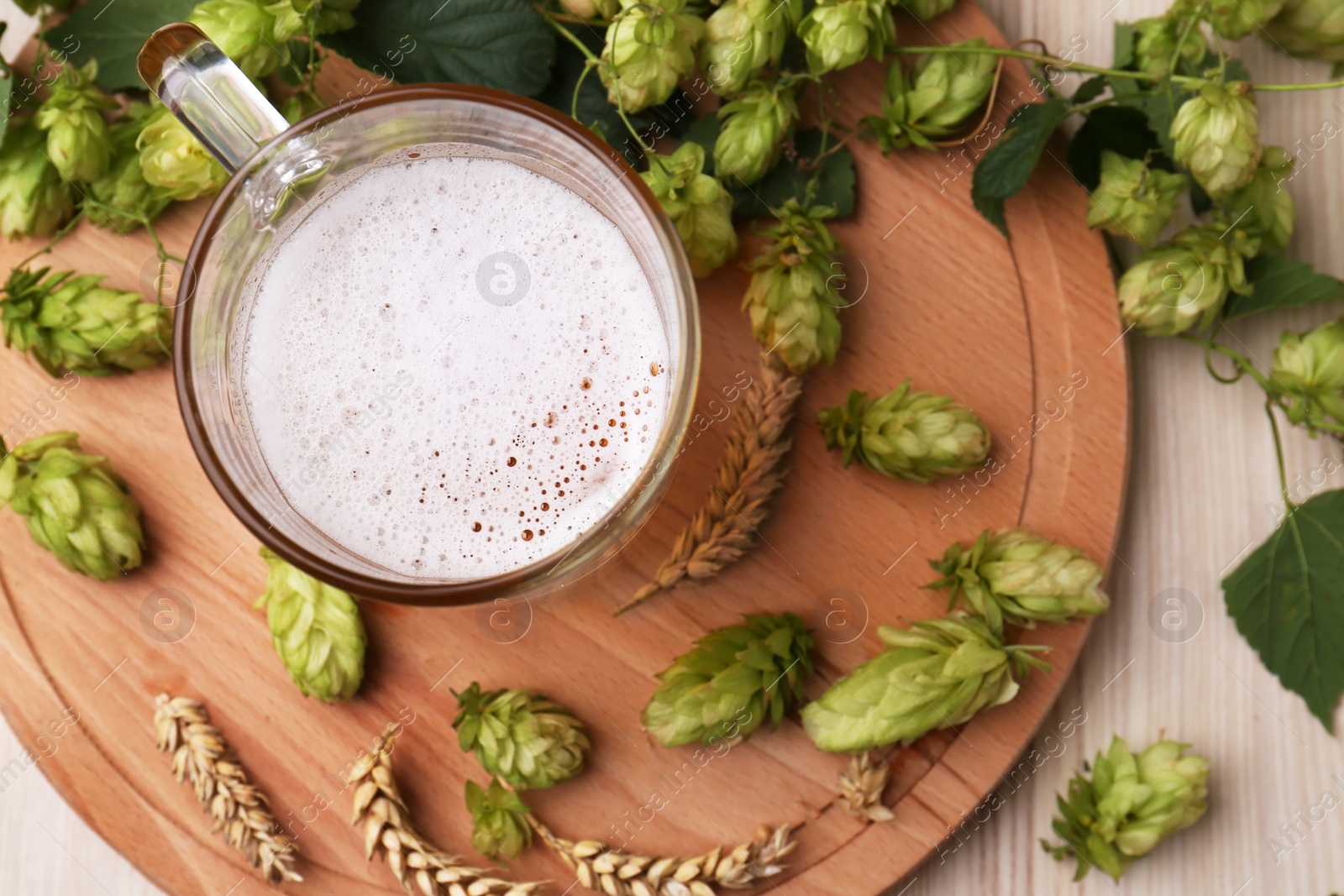 Photo of Mug with beer, fresh hops and ears of wheat on light wooden table, top view