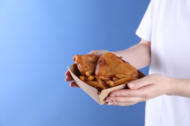 Woman holding fish and chips in paper box on blue background, closeup. Space for text