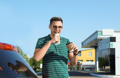 Photo of Young man with coffee eating hot dog near car at gas station