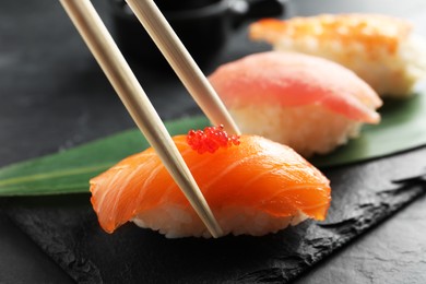Photo of Taking delicious nigiri sushi with chopsticks from serving board on black table, closeup