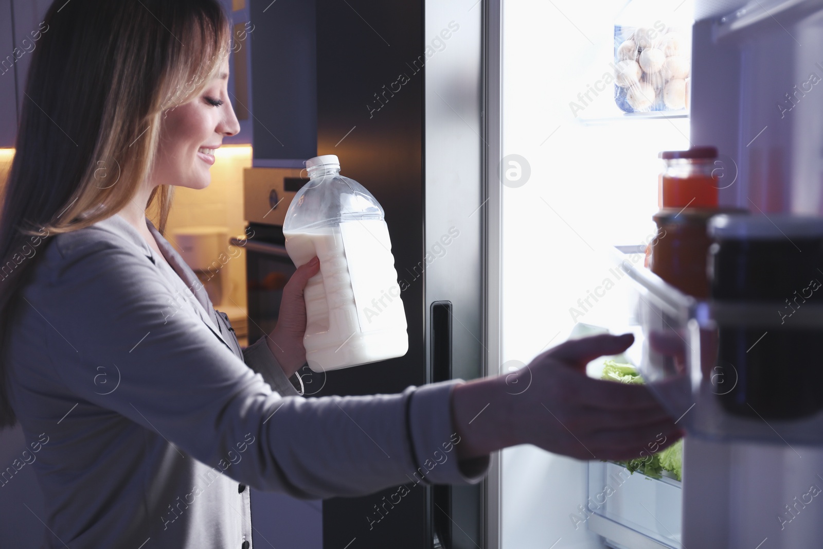 Photo of Young woman holding gallon bottle of milk near refrigerator in kitchen at night