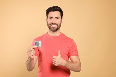 Happy man with condom showing thumb up on beige background. Safe sex