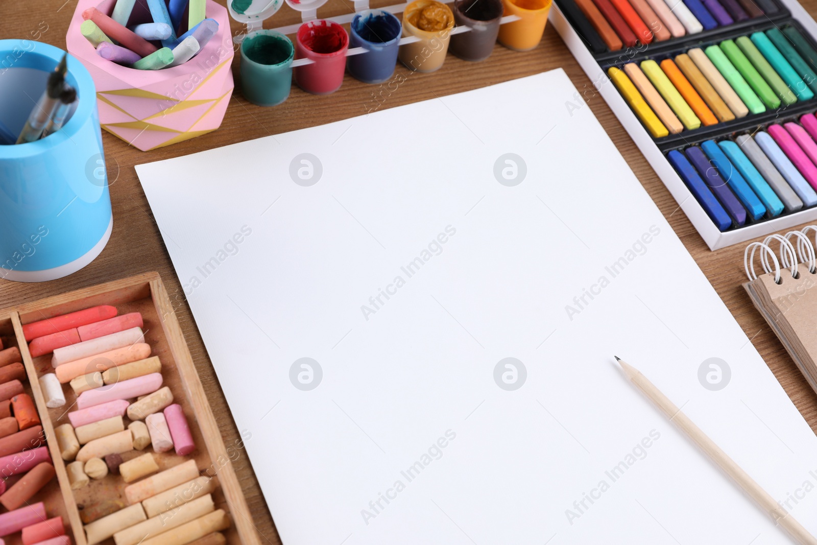 Photo of Blank sheet of paper, colorful chalk pastels and other drawing tools on wooden table. Modern artist's workplace
