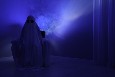 Photo of Creepy ghost. Woman covered with sheet sitting in armchair in blue light, space for text