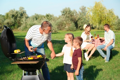 Photo of Grandfather with little kids cooking food on barbecue grill and their family in park