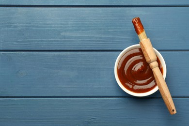 Photo of Tasty barbeque sauce in bowl and brush on blue wooden table, top view. Space for text