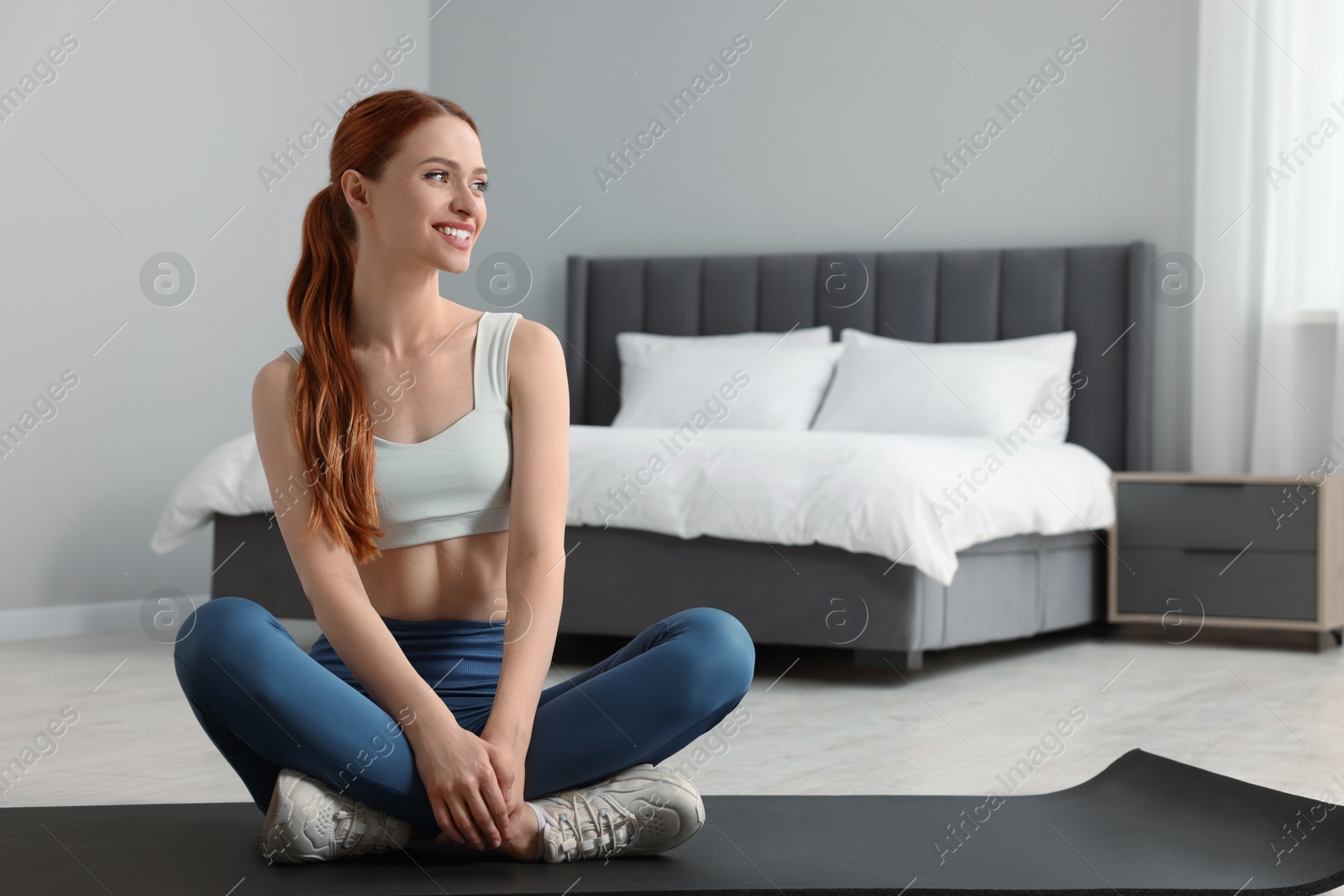 Photo of Young woman in sportswear sitting on fitness mat in bedroom, space for text