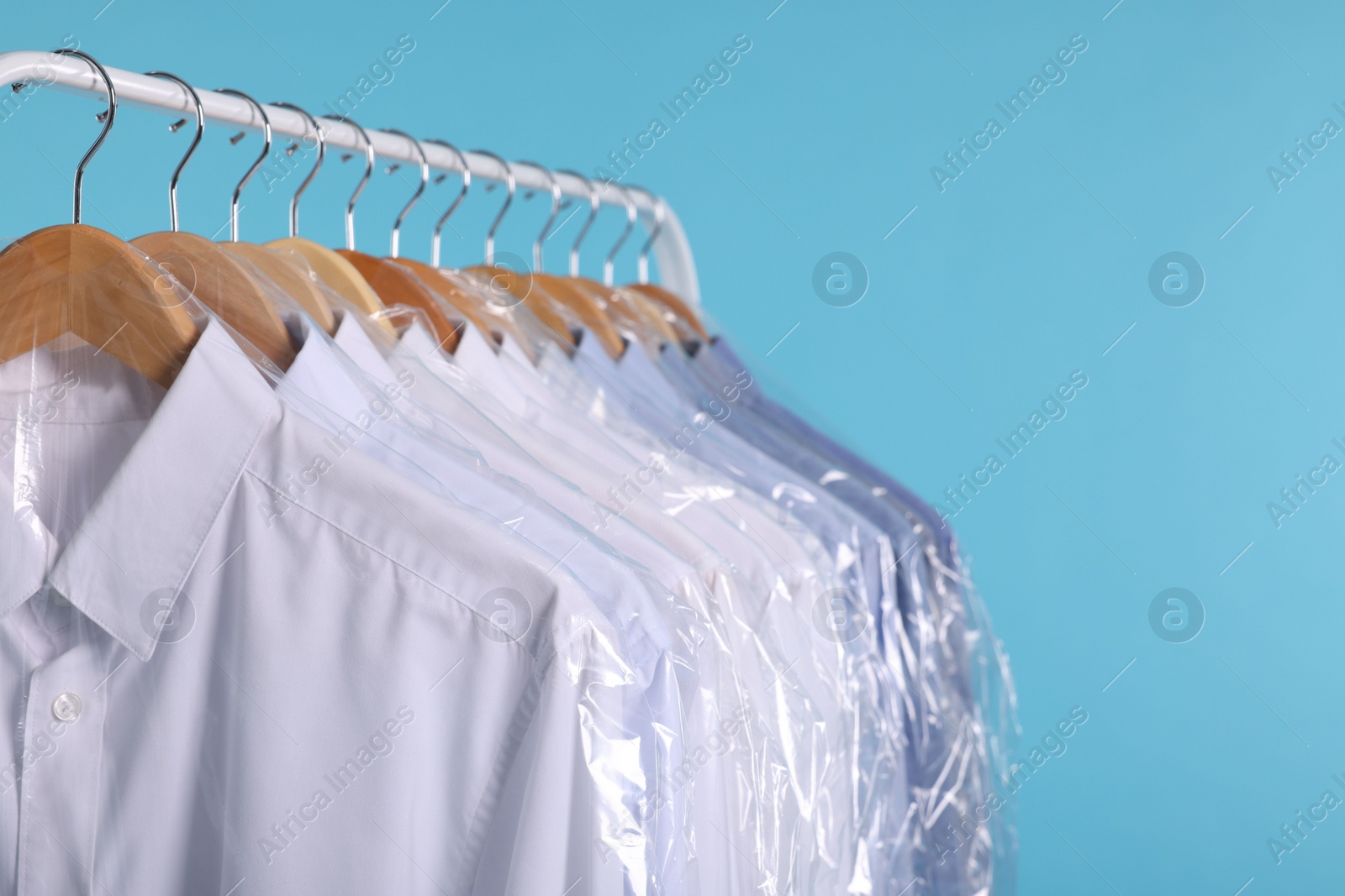 Photo of Dry-cleaning service. Many different clothes in plastic bags hanging on rack against light blue background, closeup and space for text