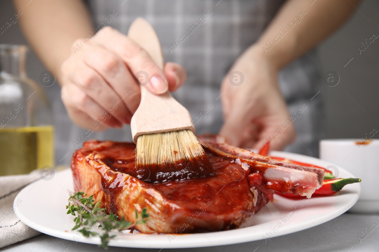 Photo of Woman spreading marinade onto raw meat with basting brush at table, closeup