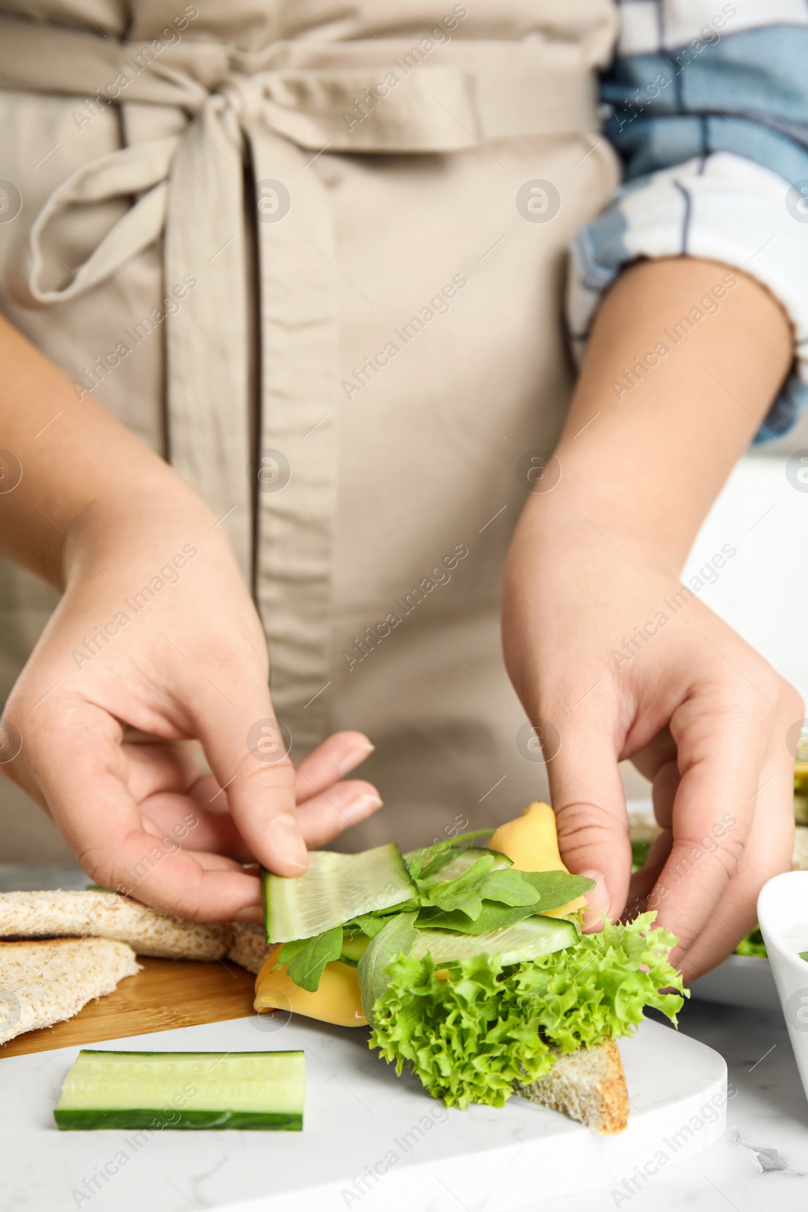 Photo of Woman adding cucumber to tasty sandwich at white marble table, closeup