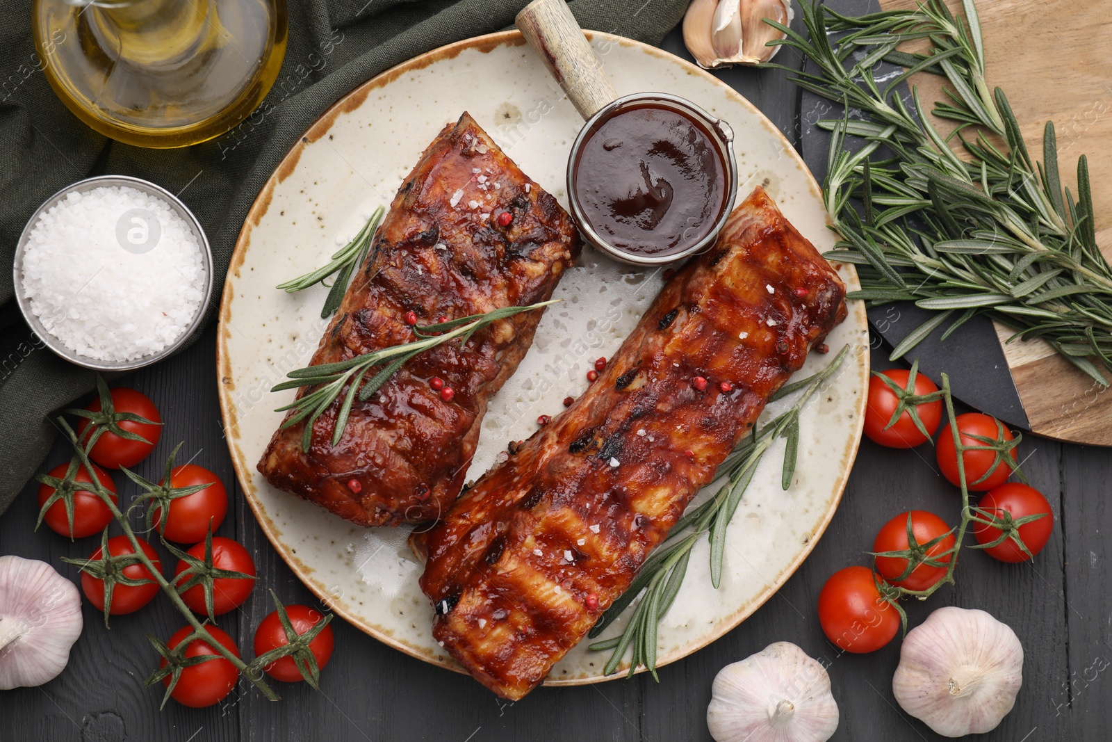 Photo of Tasty roasted pork ribs served with sauce, tomatoes and other products on grey wooden table, flat lay