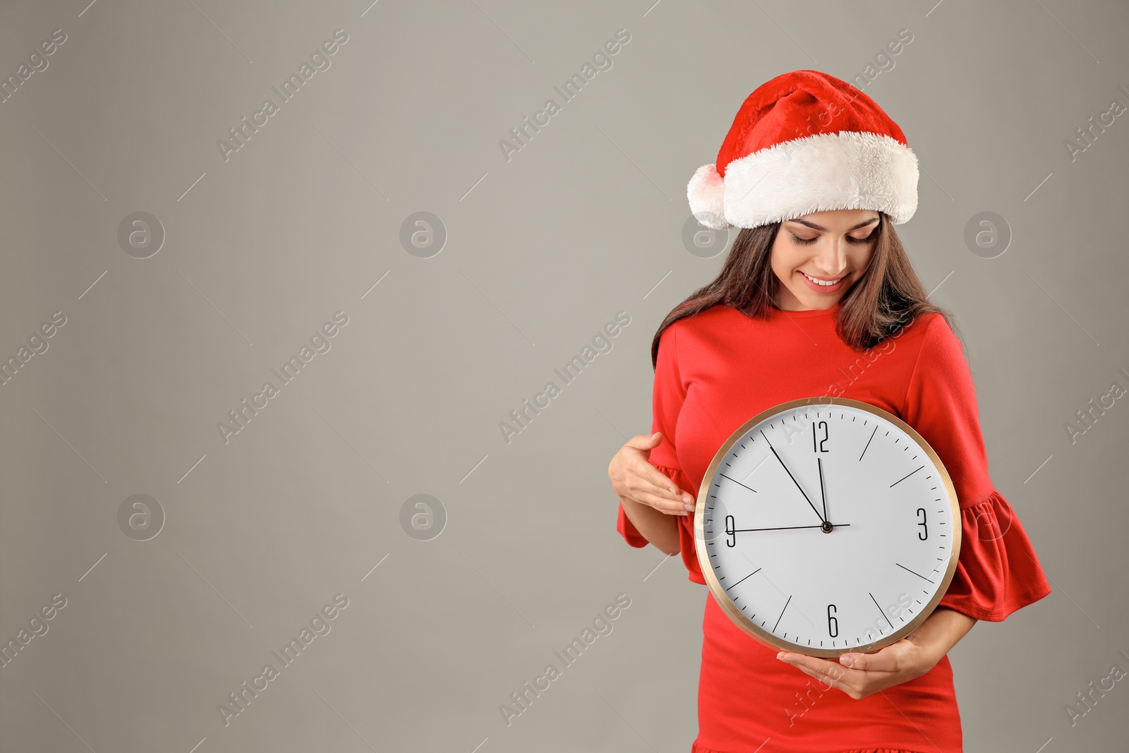 Photo of Young beautiful woman in Santa hat holding big clock on grey background. Christmas celebration