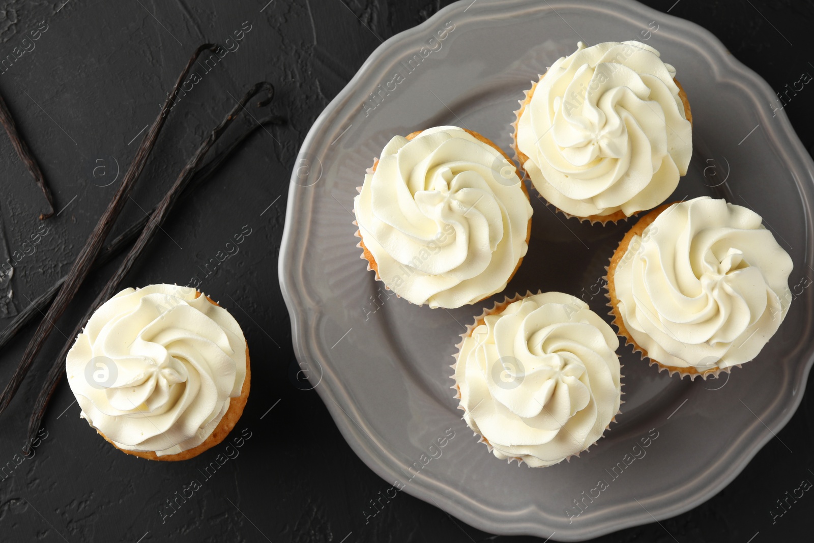 Photo of Tasty cupcakes with cream and vanilla pods on black table, top view