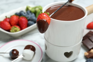 Fondue pot of melted chocolate and fork with strawberry on white marble table, closeup