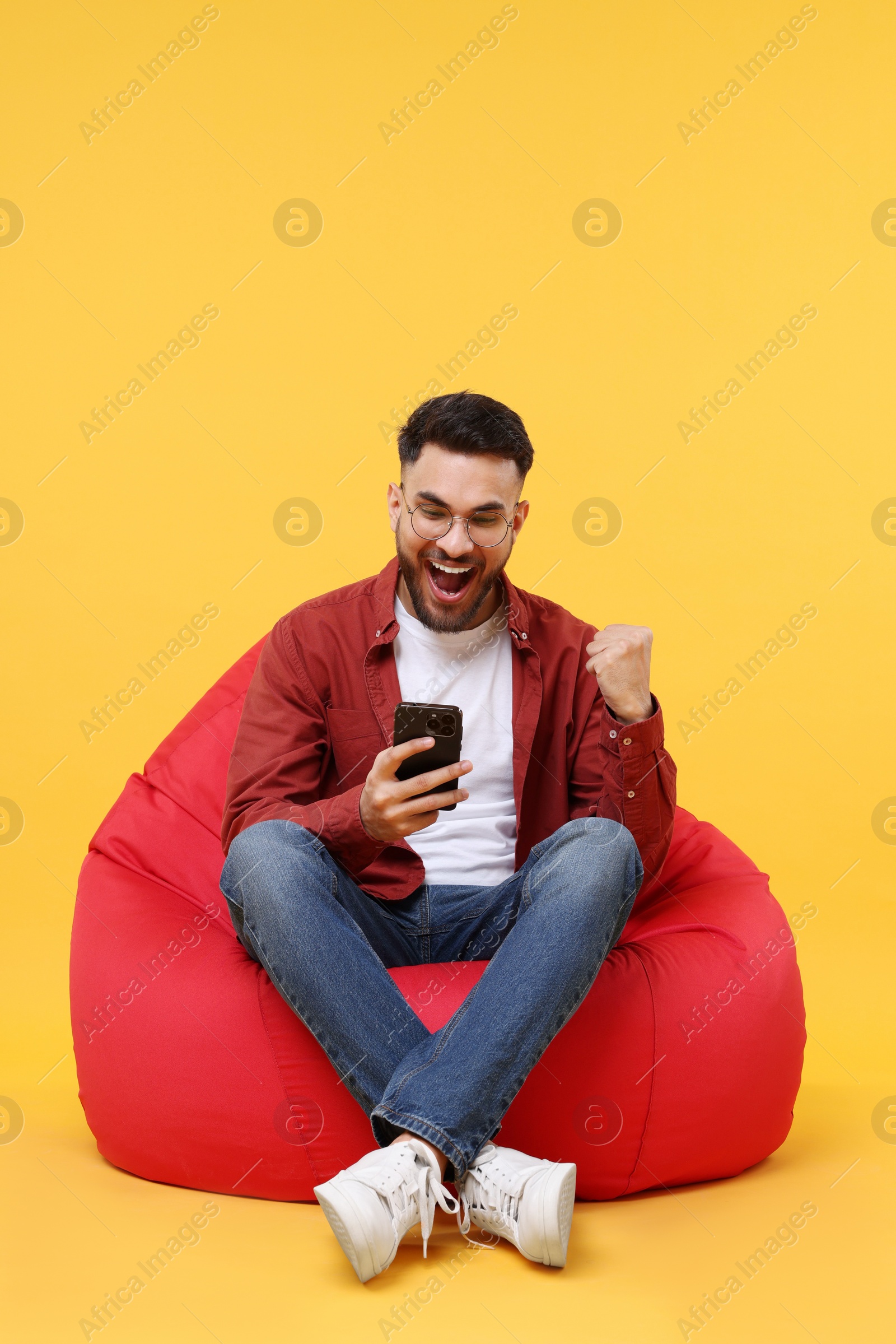 Photo of Happy young man using smartphone on bean bag chair against yellow background