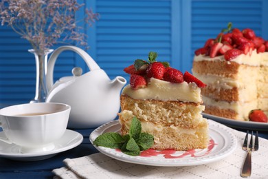 Photo of Piece of tasty cake with fresh strawberries, mint and cup of tea on blue wooden table