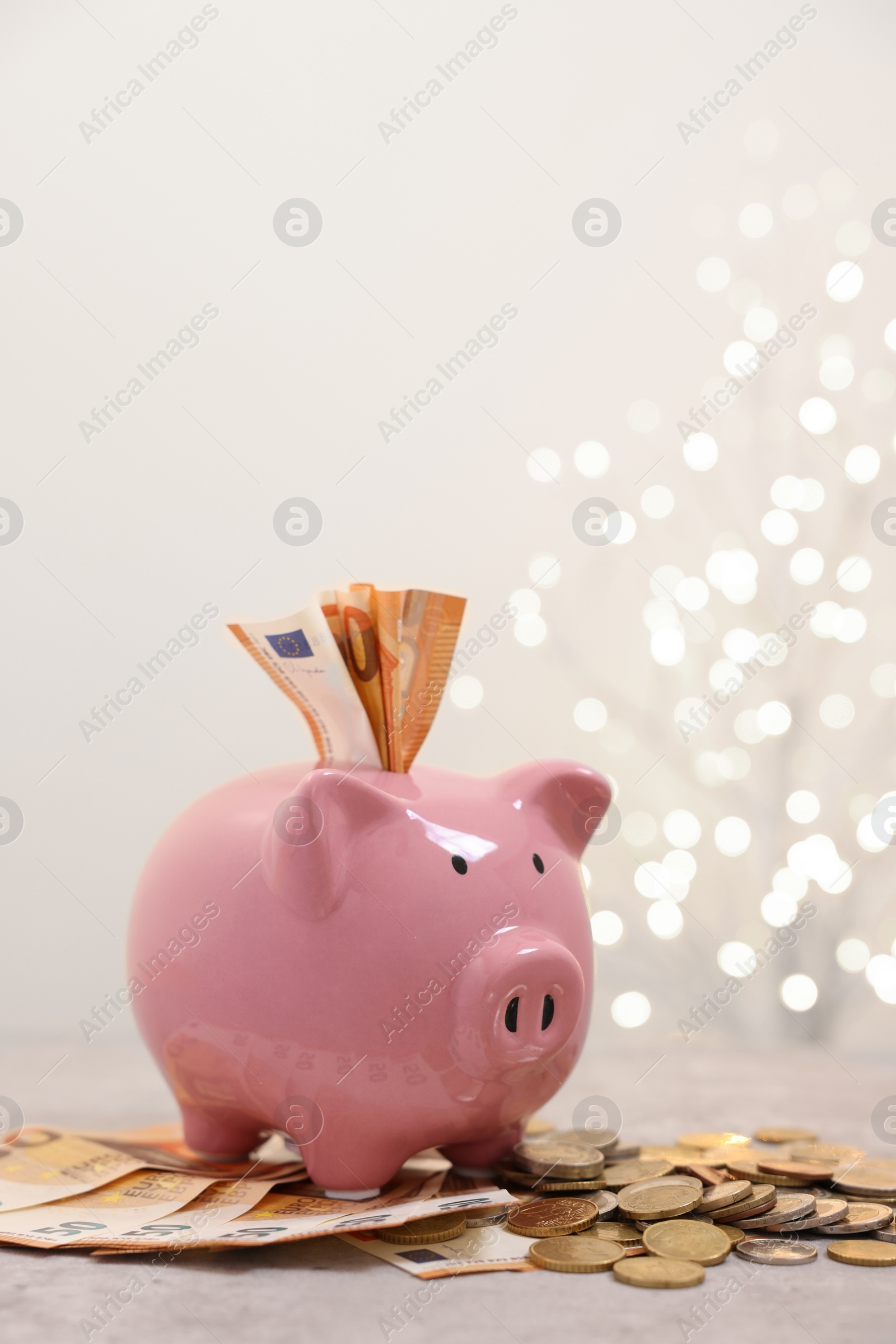 Photo of Piggy bank with euro banknotes and coins on grey table against blurred lights, space for text