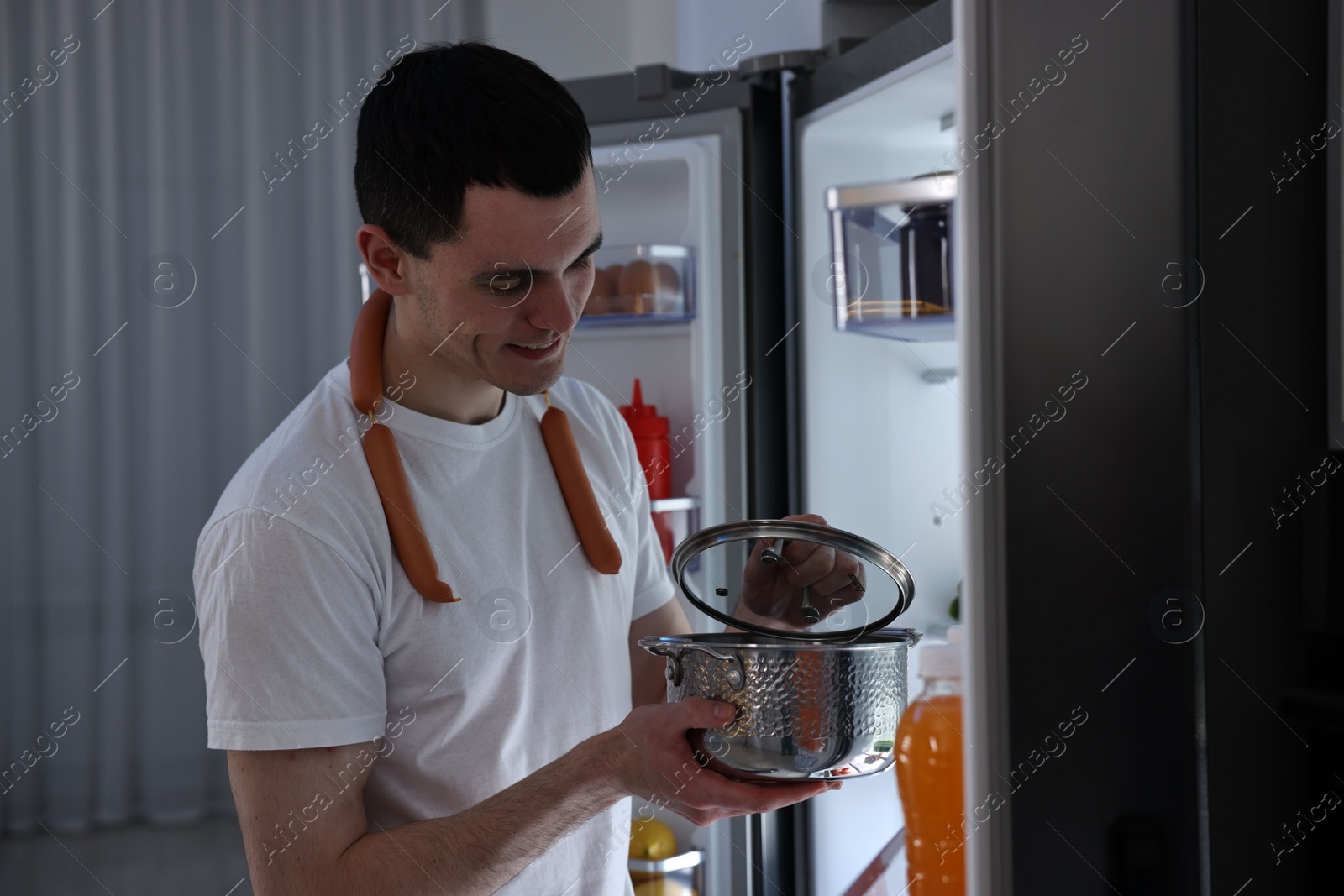 Photo of Man with sausages and pot near refrigerator in kitchen at night